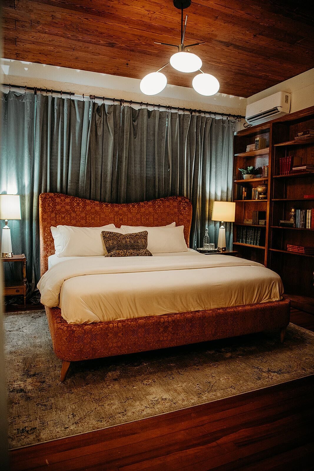 A large bed with a wooden headboard, a bedside table with a lamp, a bookshelf, and a ceiling light fixture.