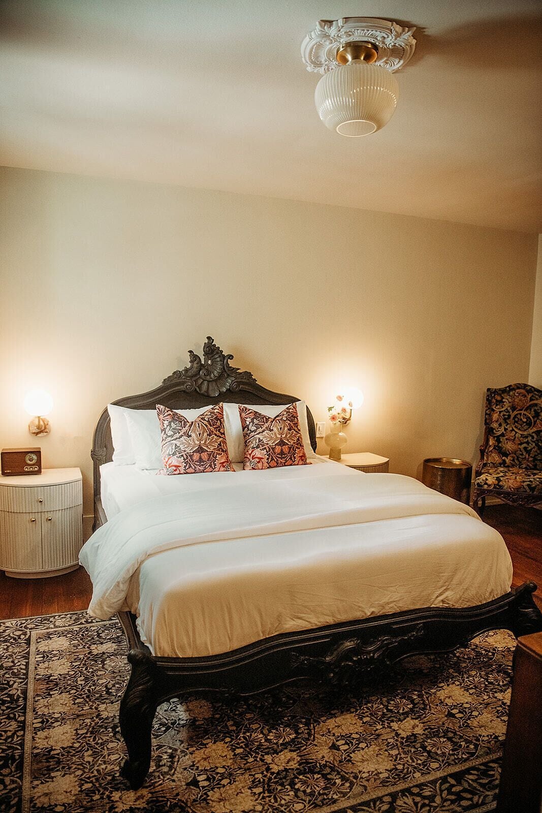 A bedroom with a large bed, ornate headboard, floral-patterned pillows, a classic chandelier, and a patterned rug.