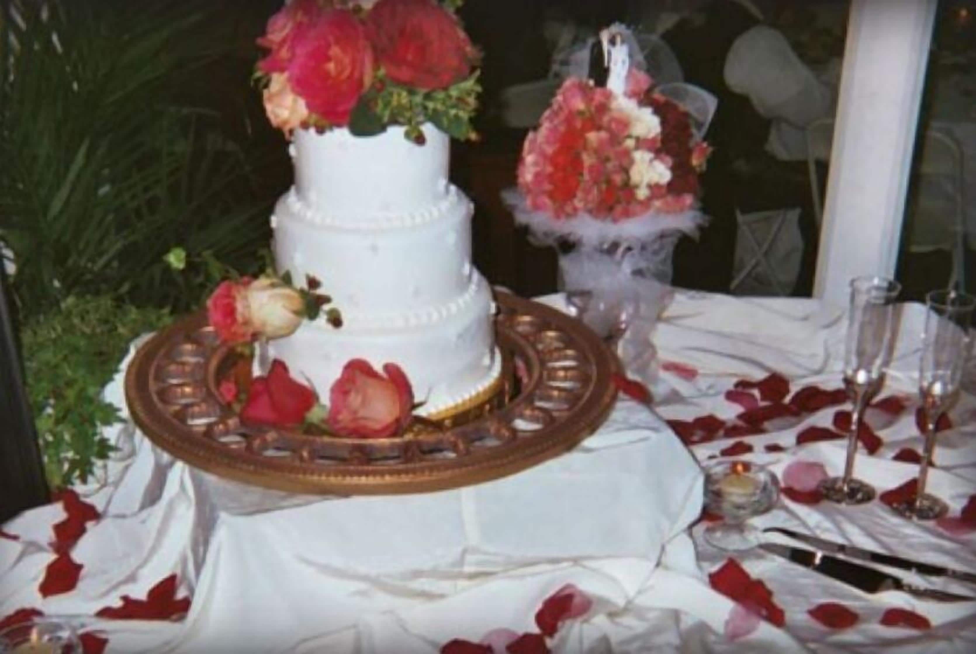 A white wedding cake with red and pink flowers on top, placed on a table with a white tablecloth and rose petals.