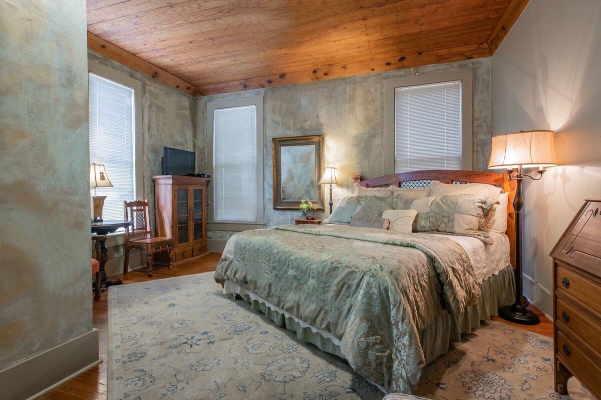 Une chambre avec un plafond en bois, un lit recouvert d’une couvre-lit à motifs floraux, une table de chevet avec une lampe et une commode.