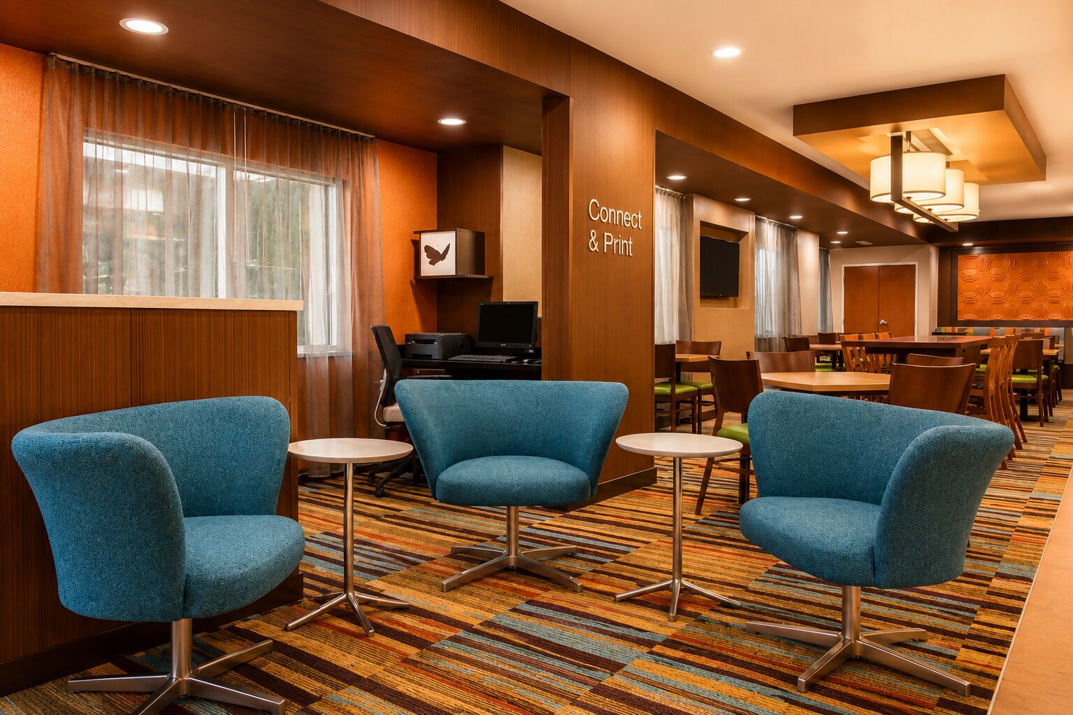 A hotel room with a desk, two blue armchairs, a small table, a computer, and a sign that reads 'Connect & Print'.