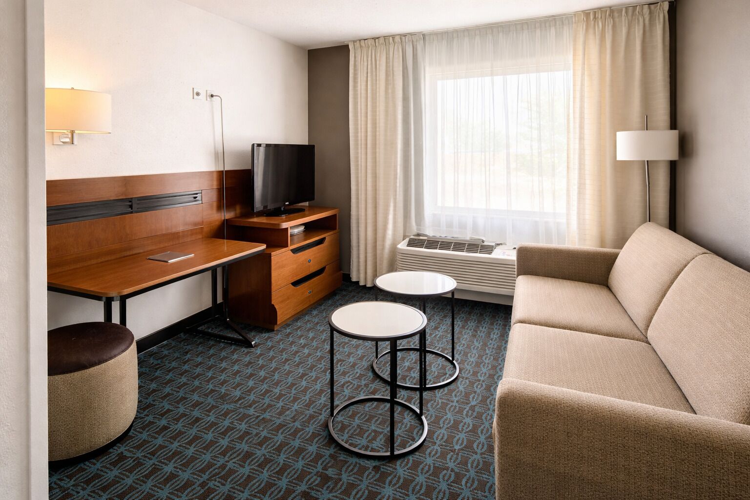A hotel room with a wooden desk, a television, a sofa, two small tables, and a window with curtains.