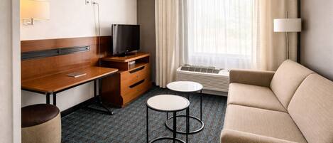 A hotel room with a wooden desk, a television, a sofa, two small tables, and a window with curtains.