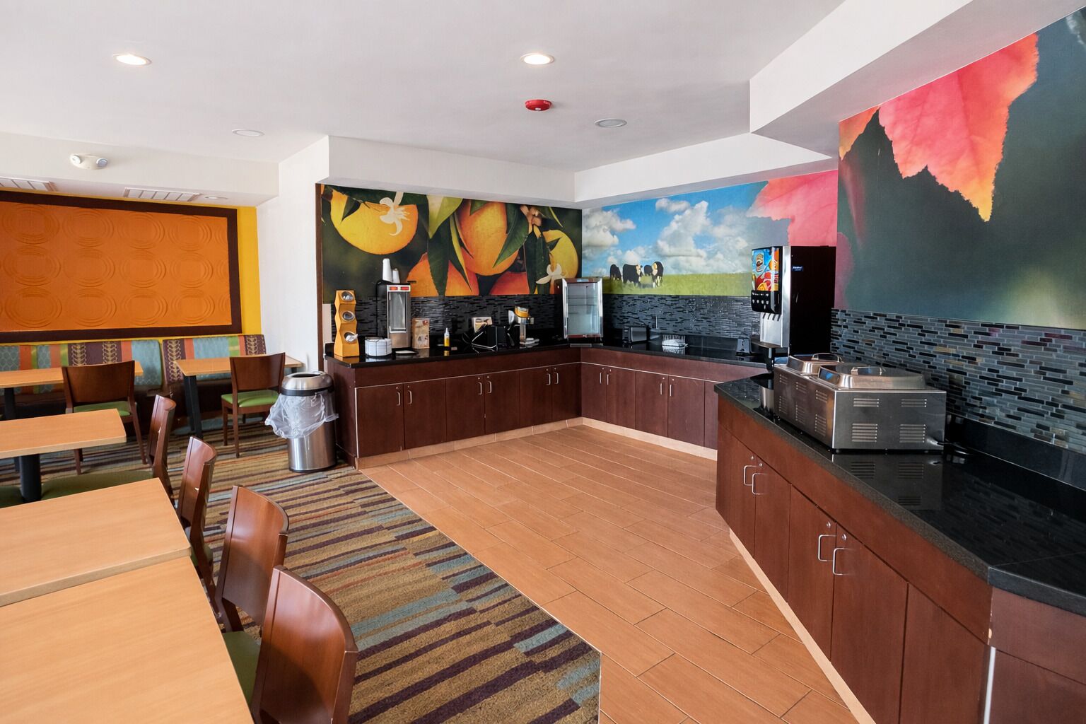 A modern kitchen with a central island, a dining area with wooden tables and chairs, and a mural with fruit and sky.