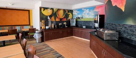 A modern kitchen with a central island, a dining area with wooden tables and chairs, and a mural with fruit and sky.