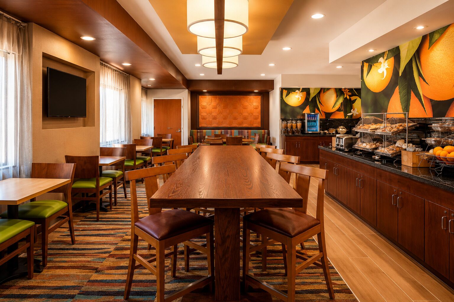 A hotel dining area with a long wooden table, multiple chairs, a buffet counter, and a large fruit-themed wall mural.