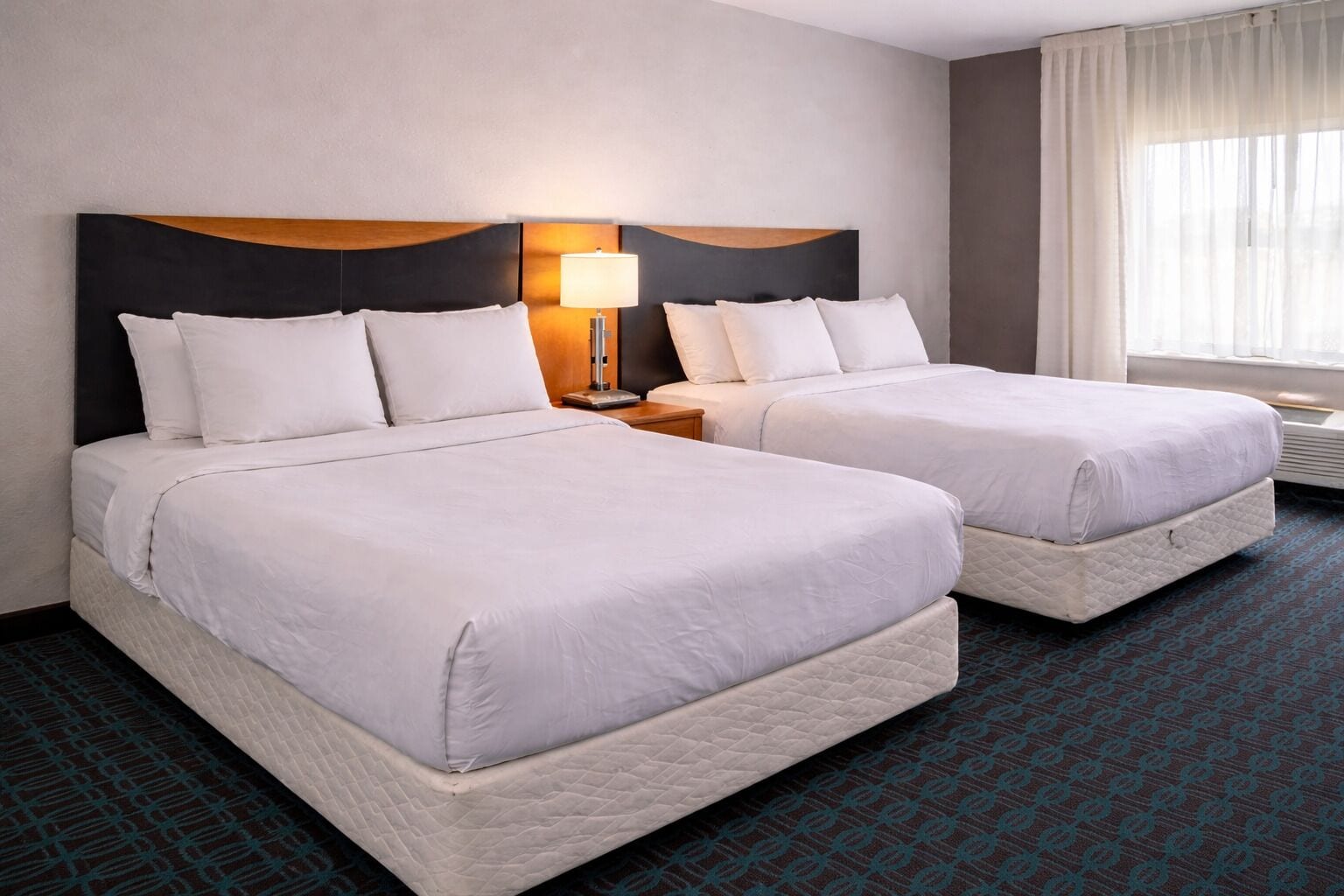 Two queen beds with white linens, a nightstand with a lamp, and a window with sheer curtains.