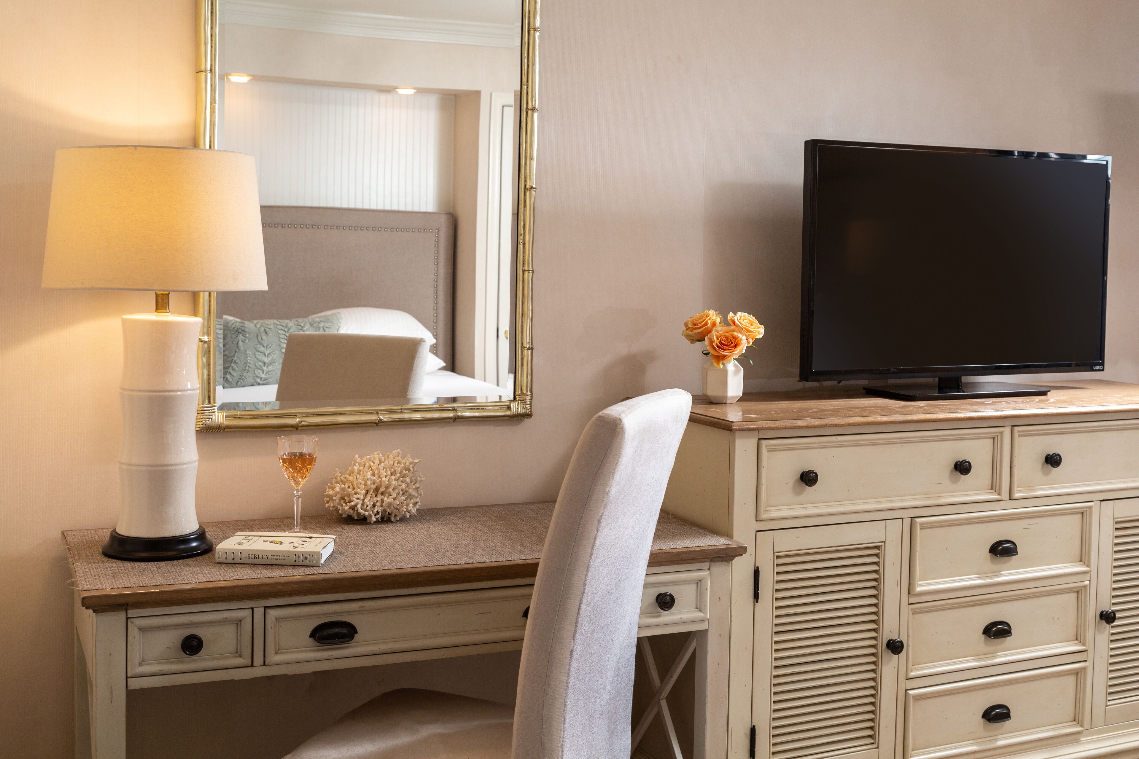 A hotel room with a desk, a television, a lamp, a vase with flowers, and a mirror.