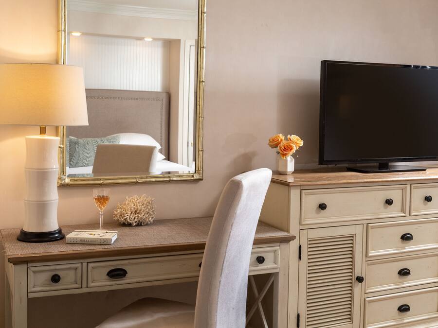 A hotel room with a desk, a television, a lamp, a vase with flowers, and a mirror.