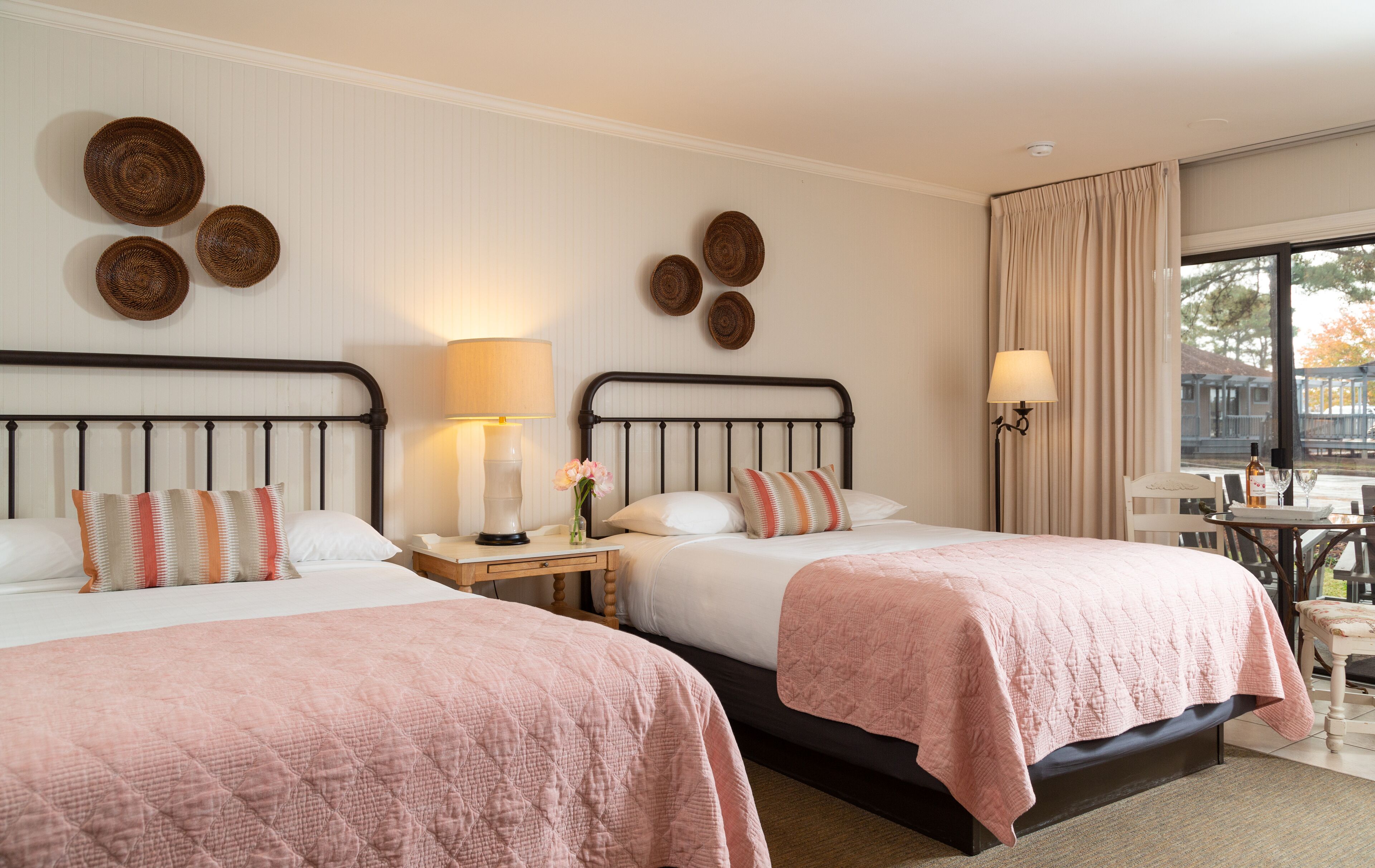 A hotel room with two beds, a nightstand with a lamp, a small table with a wine glass, and a view of the outdoors.