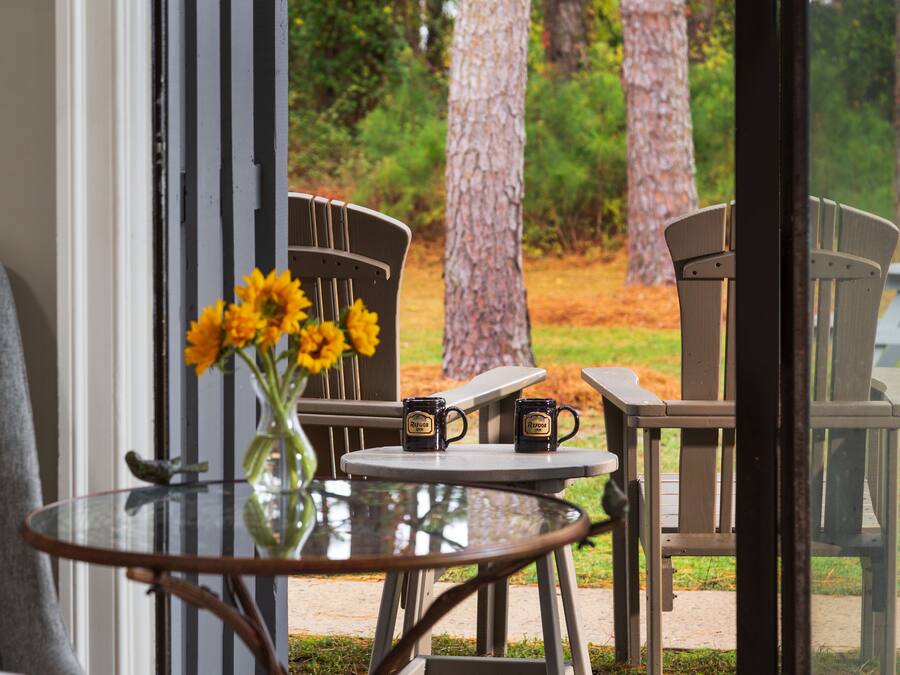 A glass table with two mugs and a vase of flowers, a gray chair, and a view of trees and grass outside.