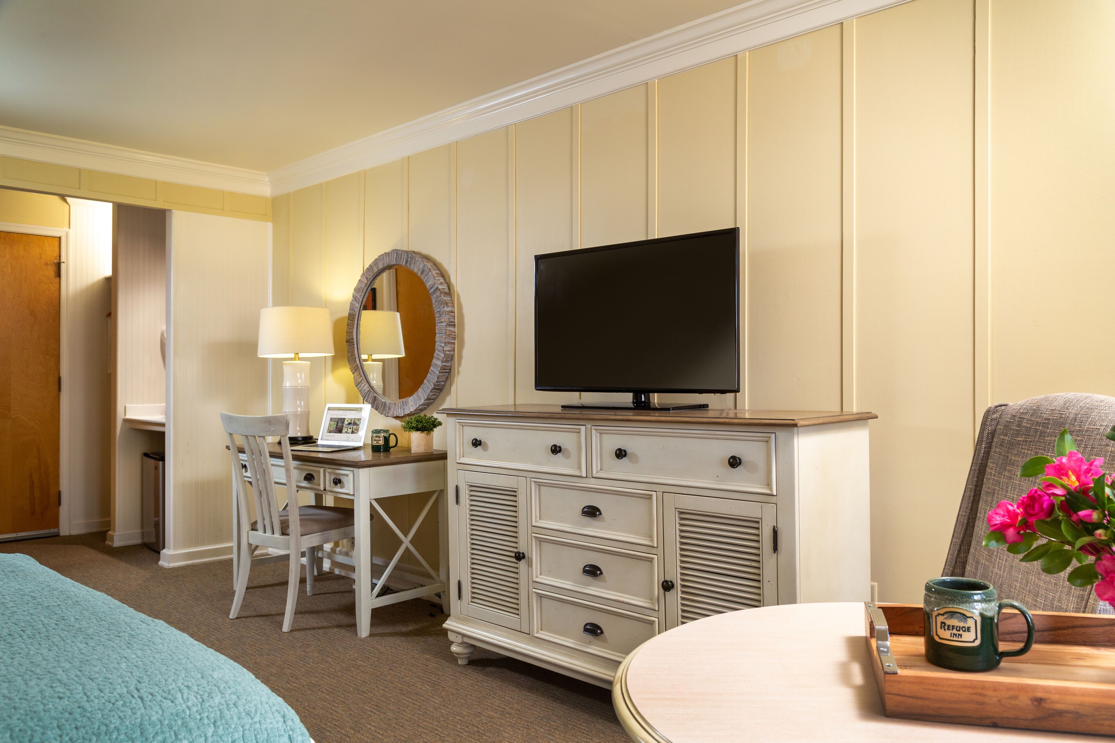 Ein Hotelzimmer mit Bett, Schreibtisch mit Lampe, Kommode mit Fernseher und Stuhl mit Blumenarrangement.