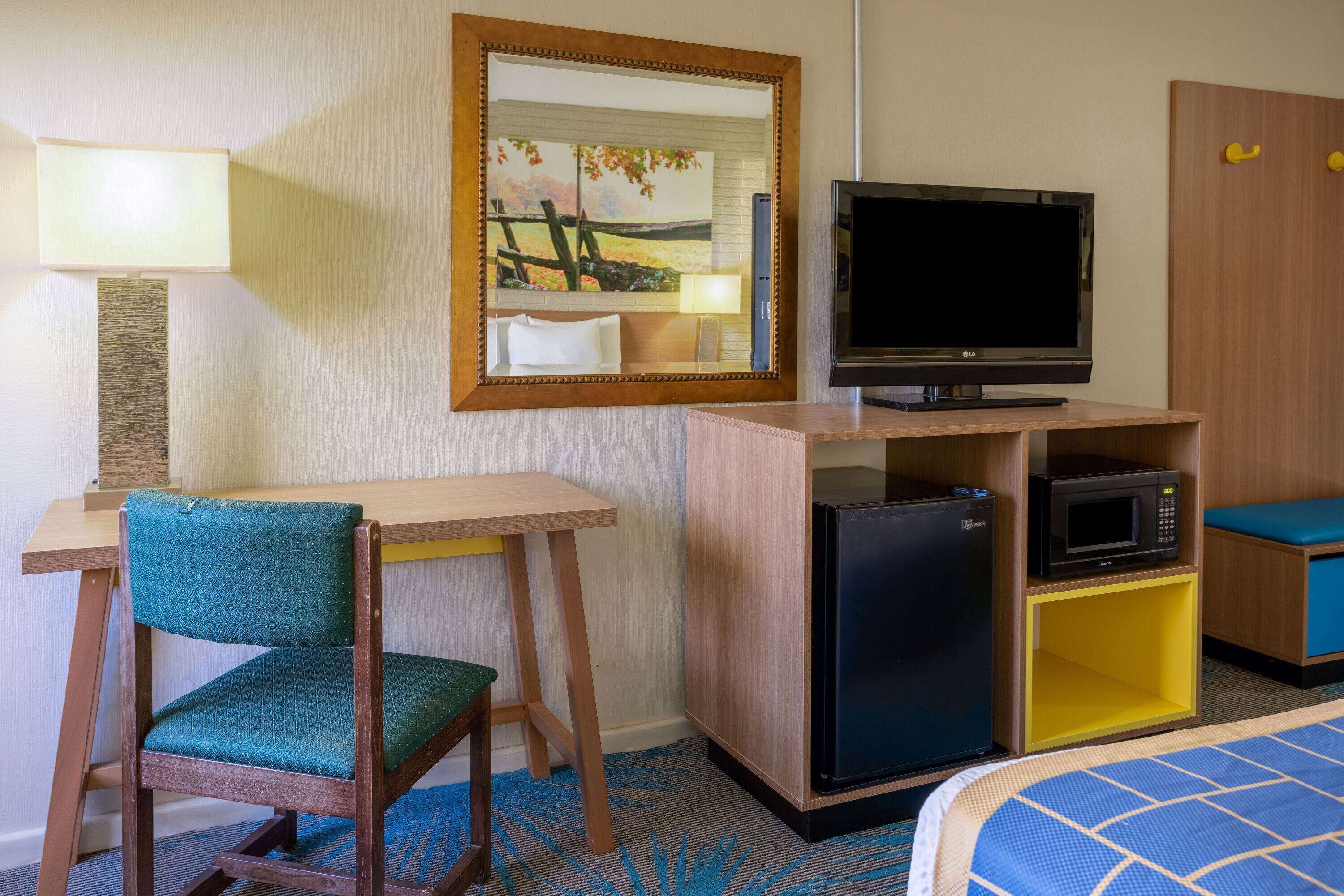 Habitación de hotel con escritorio, silla, televisión, refrigerador, microondas y cama con edredón de diseño azul y blanco.
