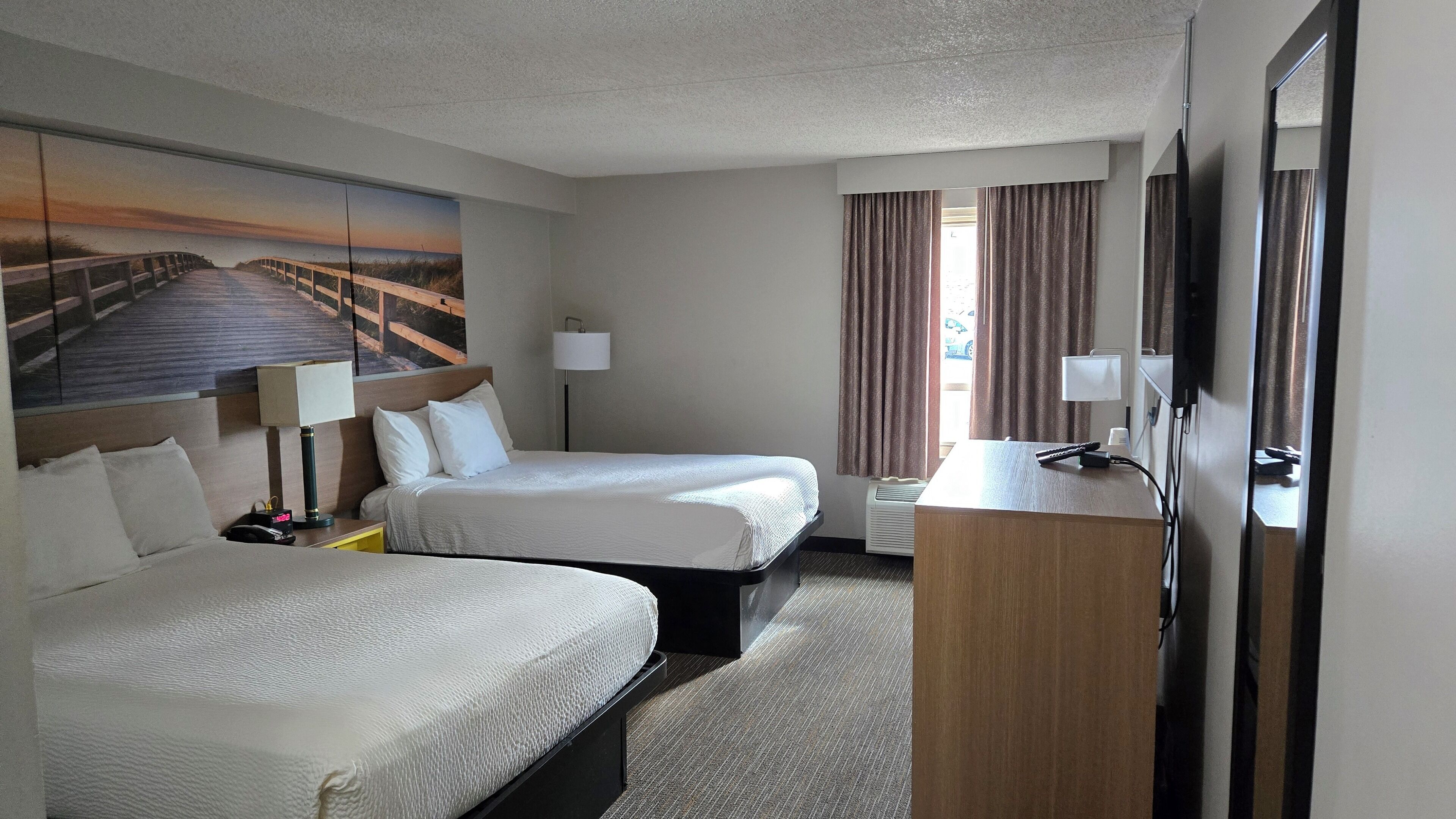 A hotel room with two beds, a desk, a TV, and a painting of a beach scene.