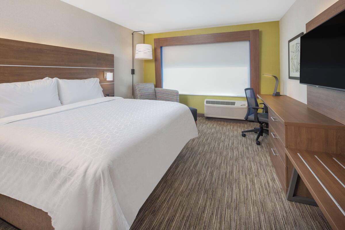 A hotel room with a large bed, a desk with a chair, a flat-screen TV, a window with blinds, and a wall-mounted lamp.