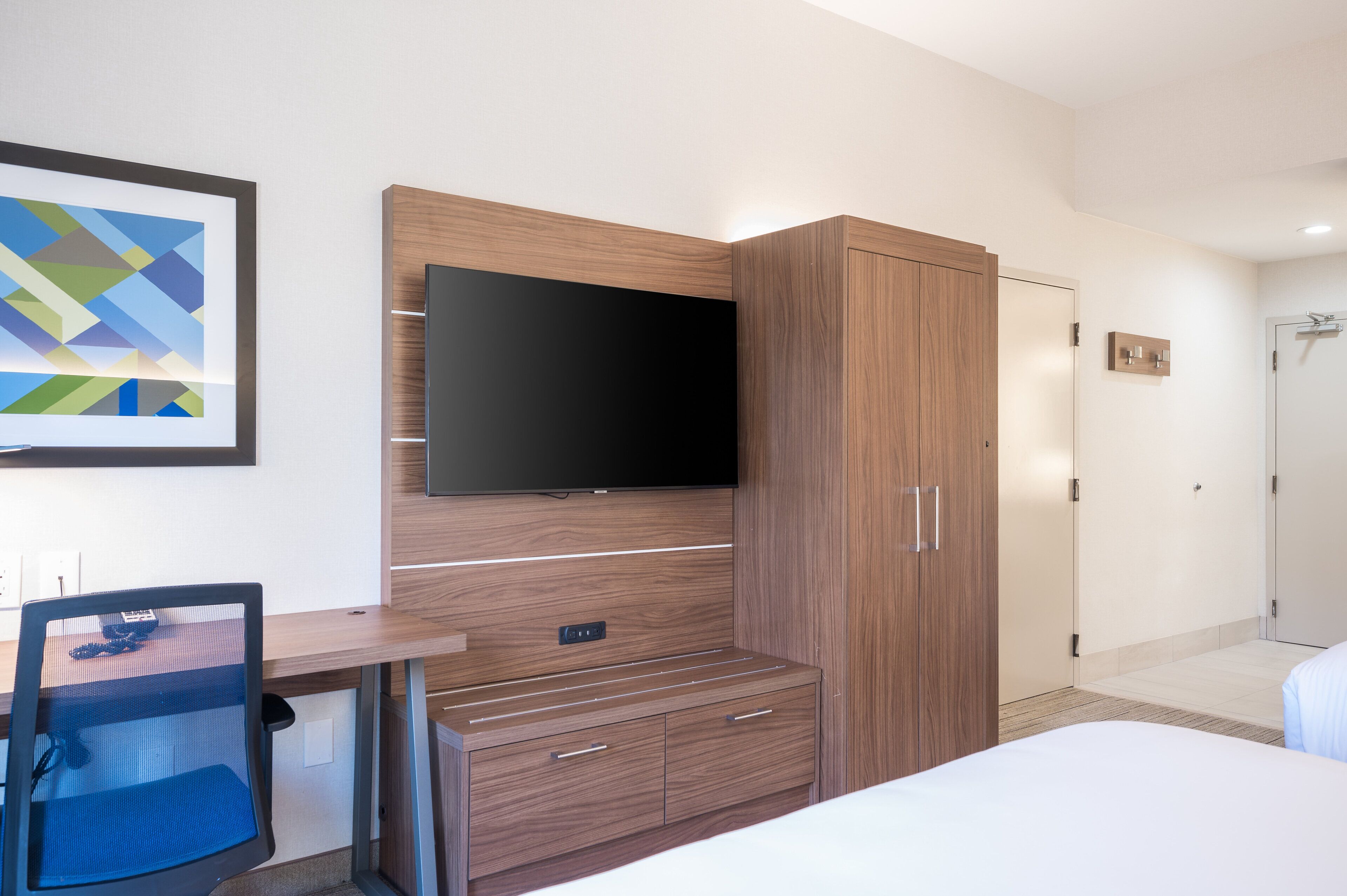 A modern hotel room with a large flat-screen TV mounted on a wooden cabinet, a desk with a chair, and a bed with white bedding.