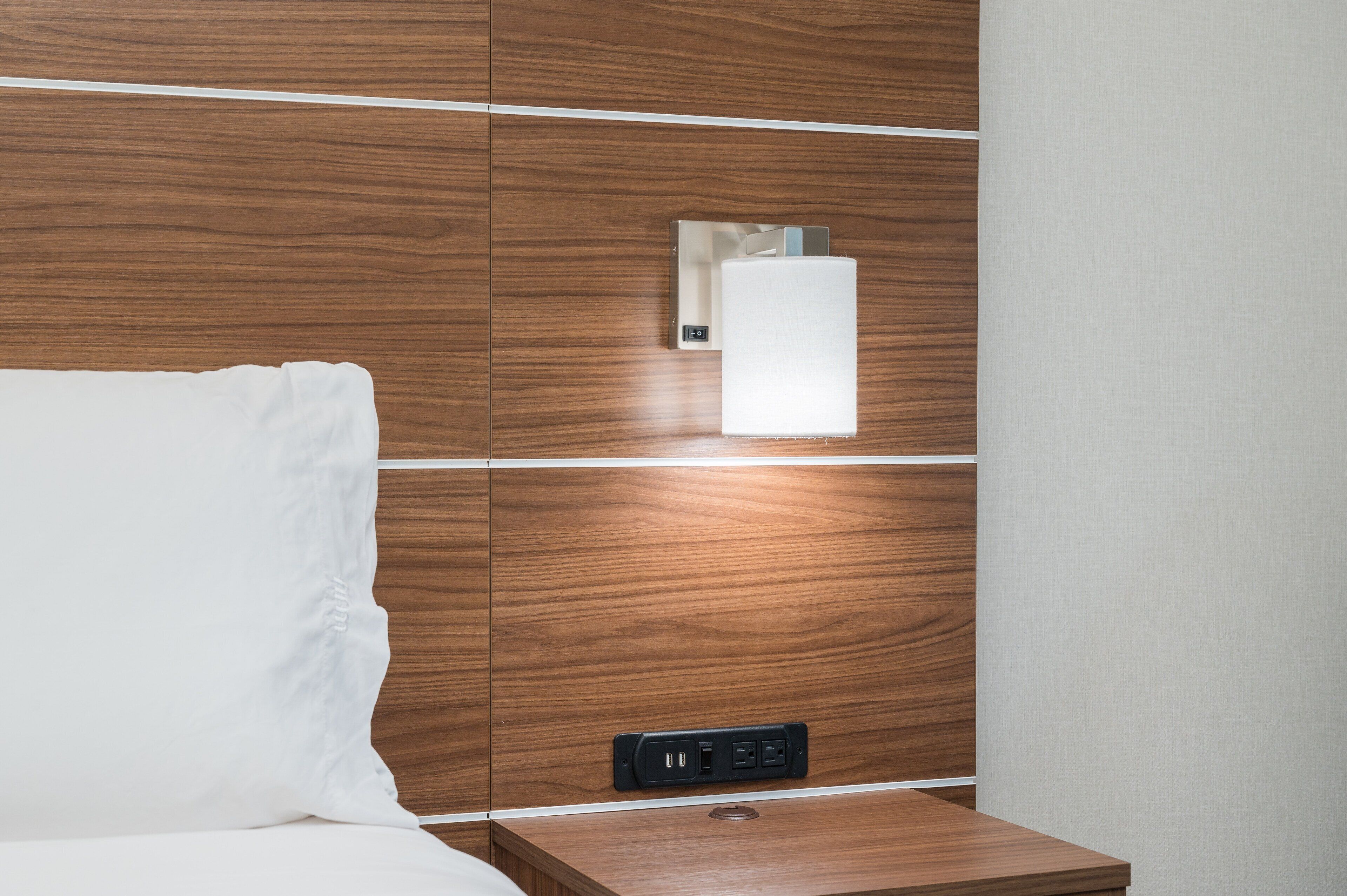 Ein Hotelzimmer mit einem Holz-Kopfteil, einem Nachttisch mit Steckdose und einer Wandleuchte.