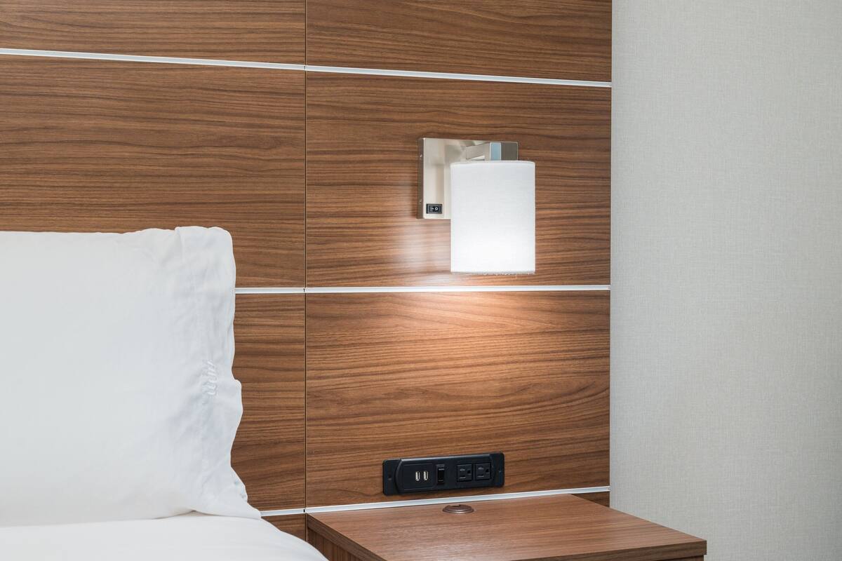 A hotel room with a wooden headboard, a bedside table with a power outlet, and a wall-mounted light fixture.