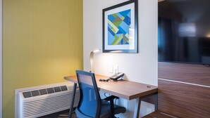A hotel room with a desk, chair, air conditioner, and a framed picture on the wall.
