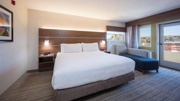 A modern hotel room with a large bed, a bedside table, a chair, and a view of the outside.