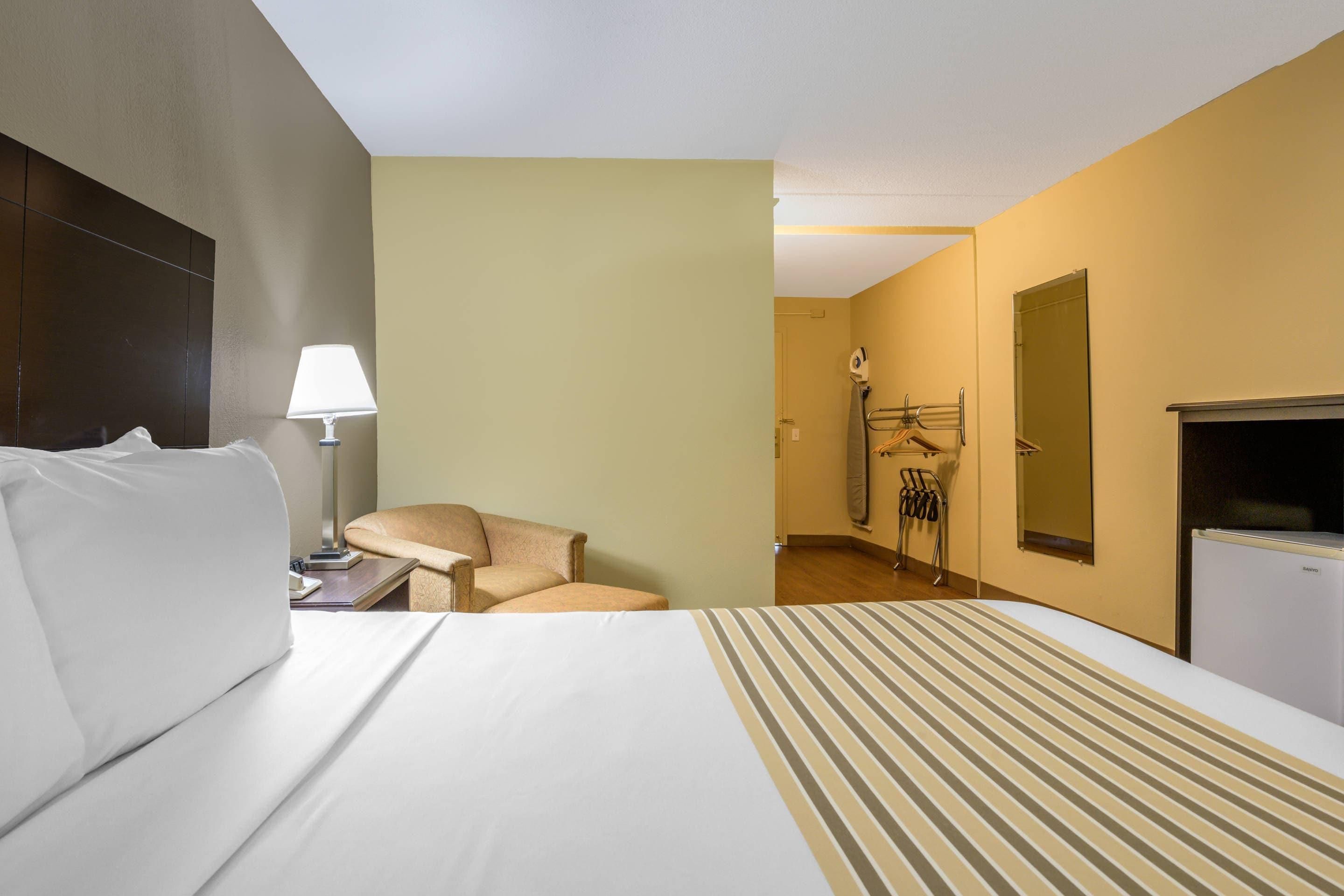 A hotel room with a bed, a chair, a desk with a lamp, a wardrobe, and a mirror.