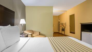 Una habitación de hotel con cama, una silla, escritorio con lámpara, armario y espejo.