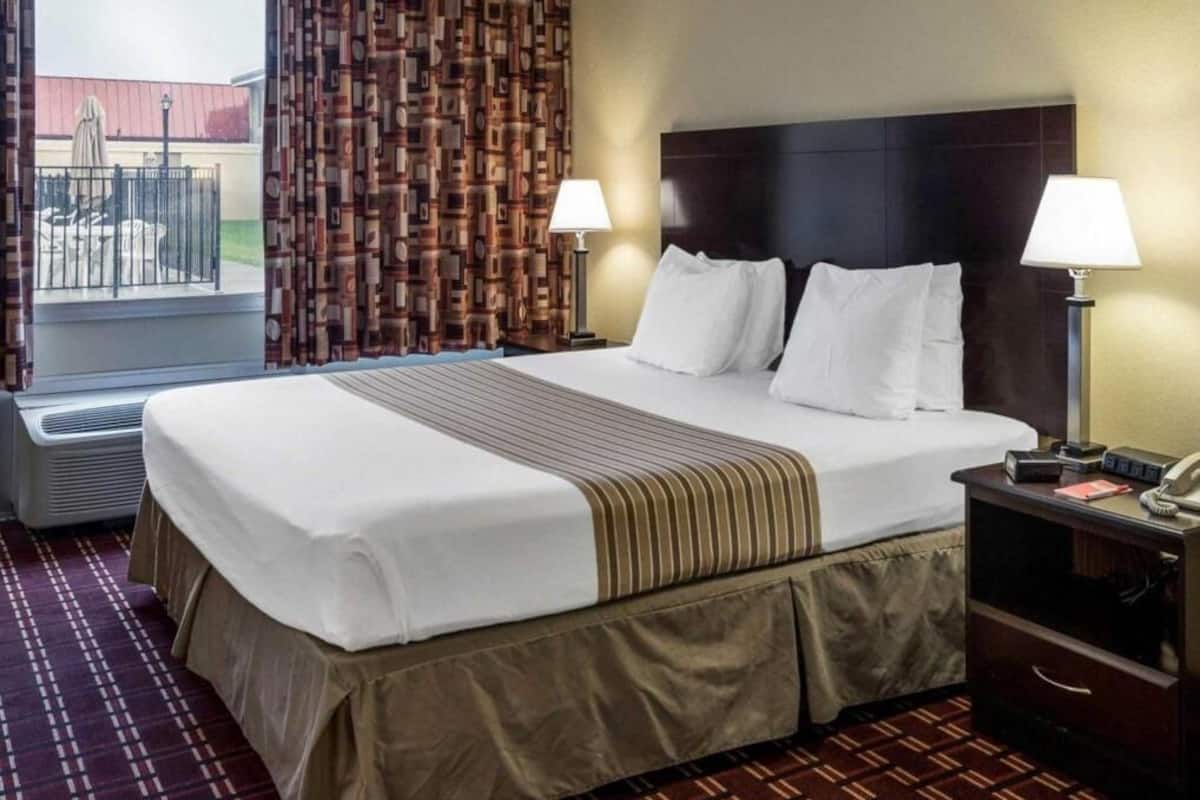 A hotel room with a large bed, two bedside lamps, a nightstand with a phone, and a view of the outside.