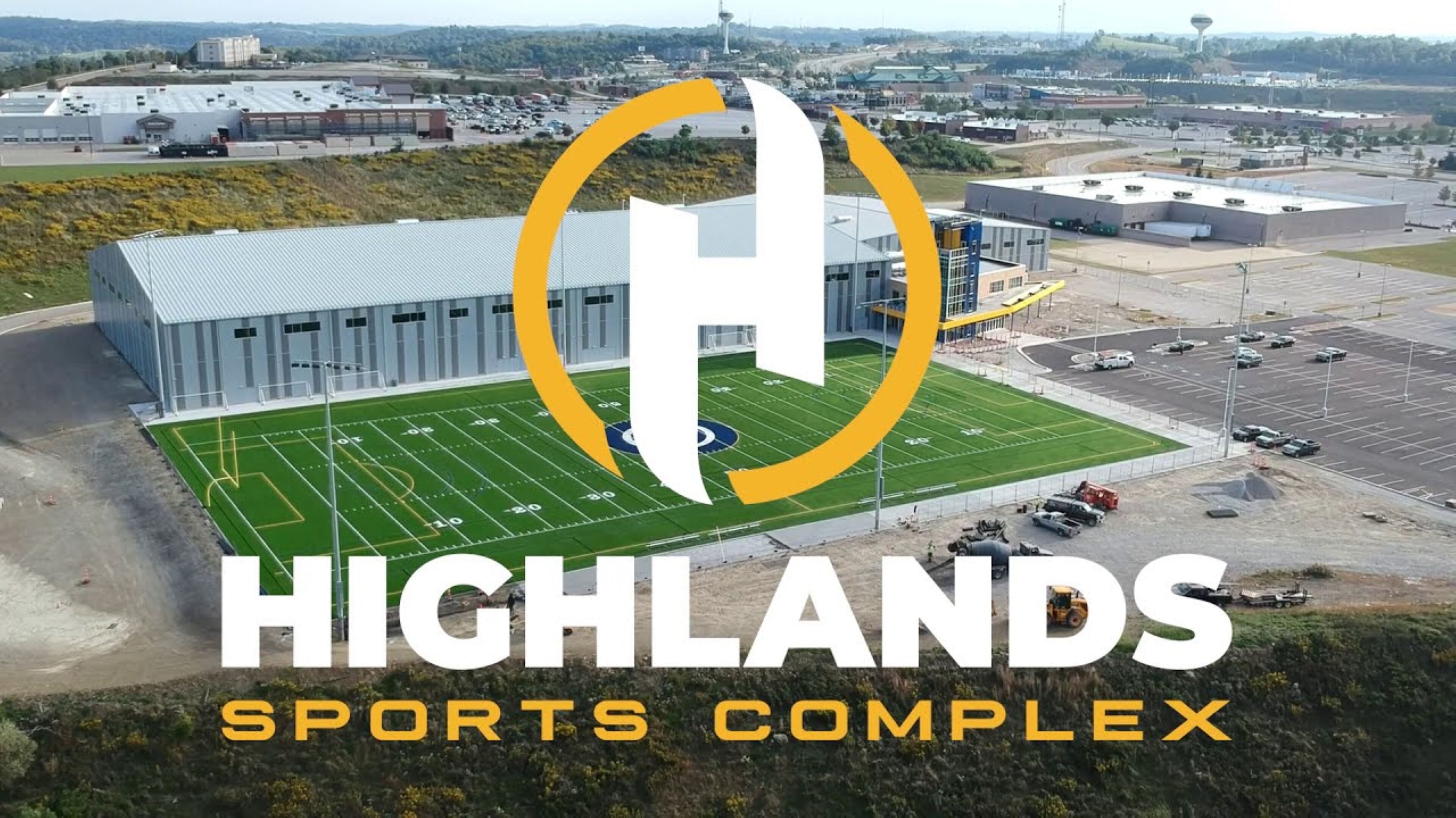 A sports complex with a large building, a football field, and a parking area.