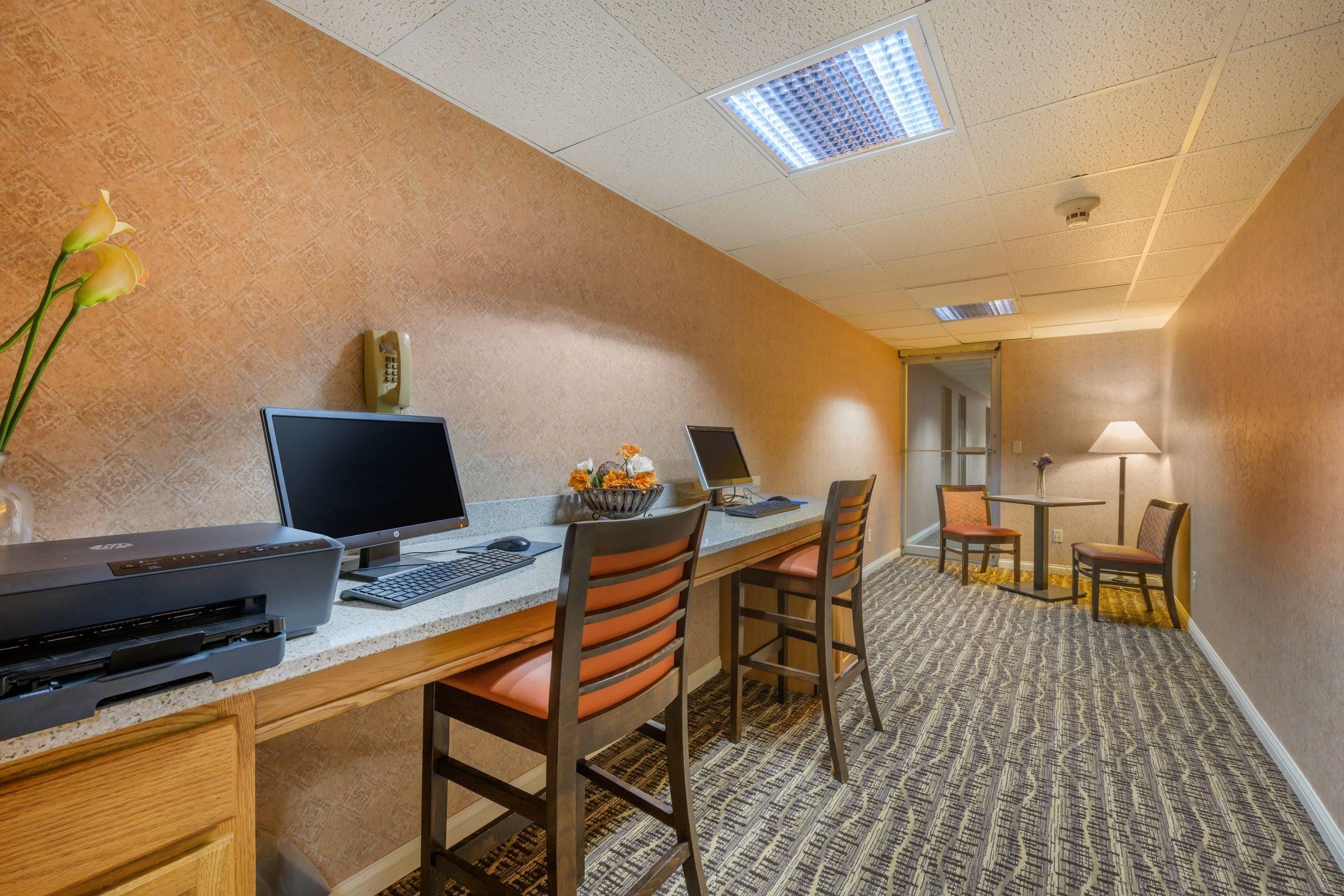 A hotel room with a desk, two chairs, a printer, a telephone, and a small table with a lamp.