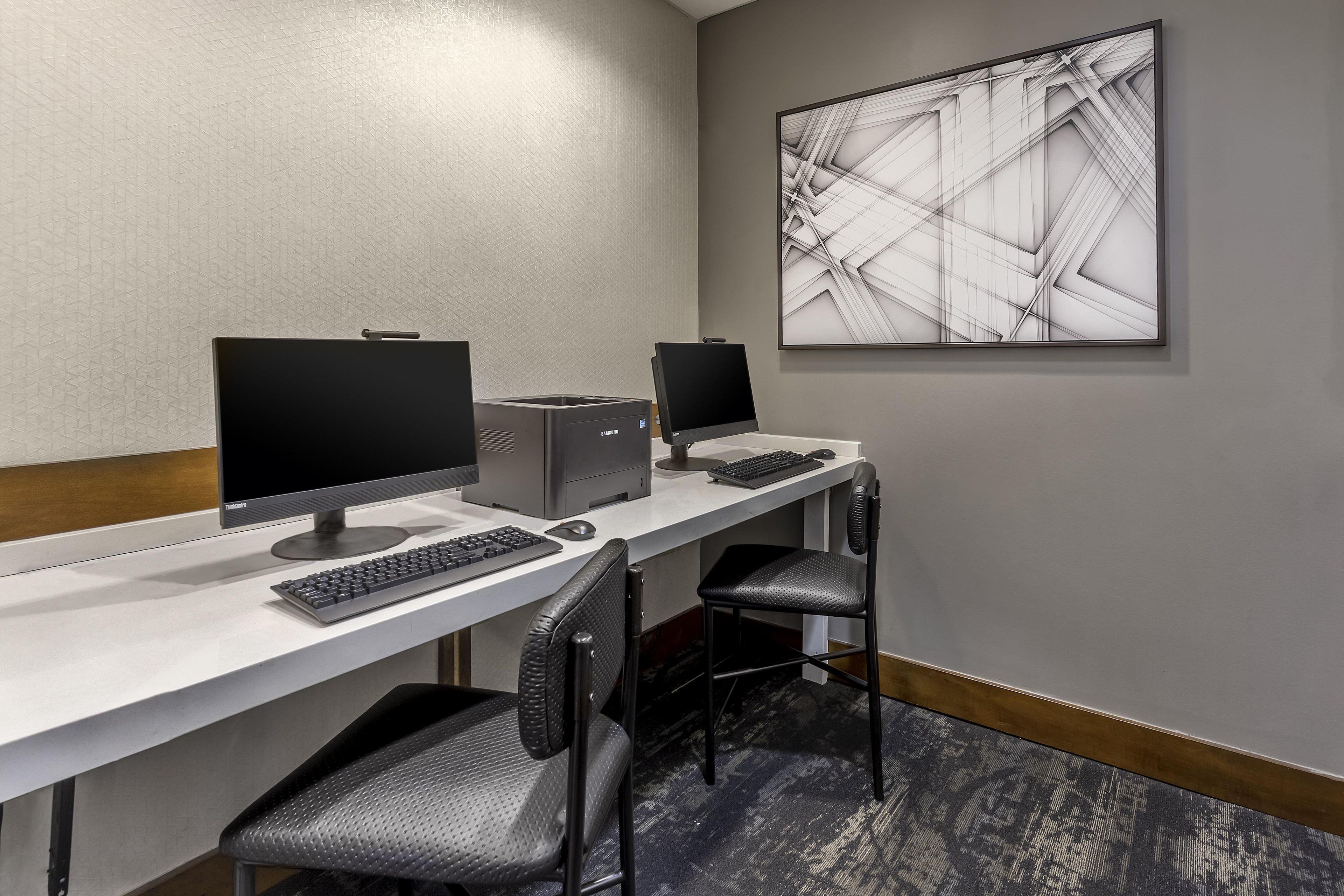 A hotel room with a desk, two monitors, a keyboard, a mouse, and a chair.