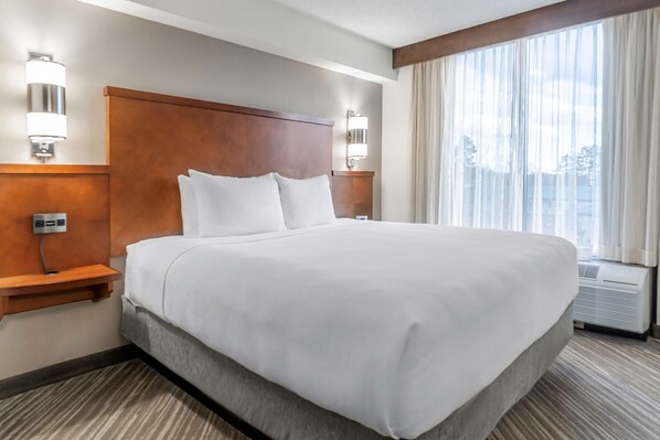 A hotel room with a large bed, two bedside tables, a wall-mounted lamp, and a large window with curtains.