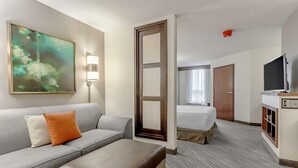 Ein Hotelzimmer mit grauem Sofa, einem braunen Hocker, einem Bett mit weißer Bettdecke, einem Fernseher und einem Bild an der Wand.