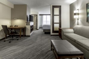 A hotel room with a desk, chair, sofa, and a small table with a lamp.