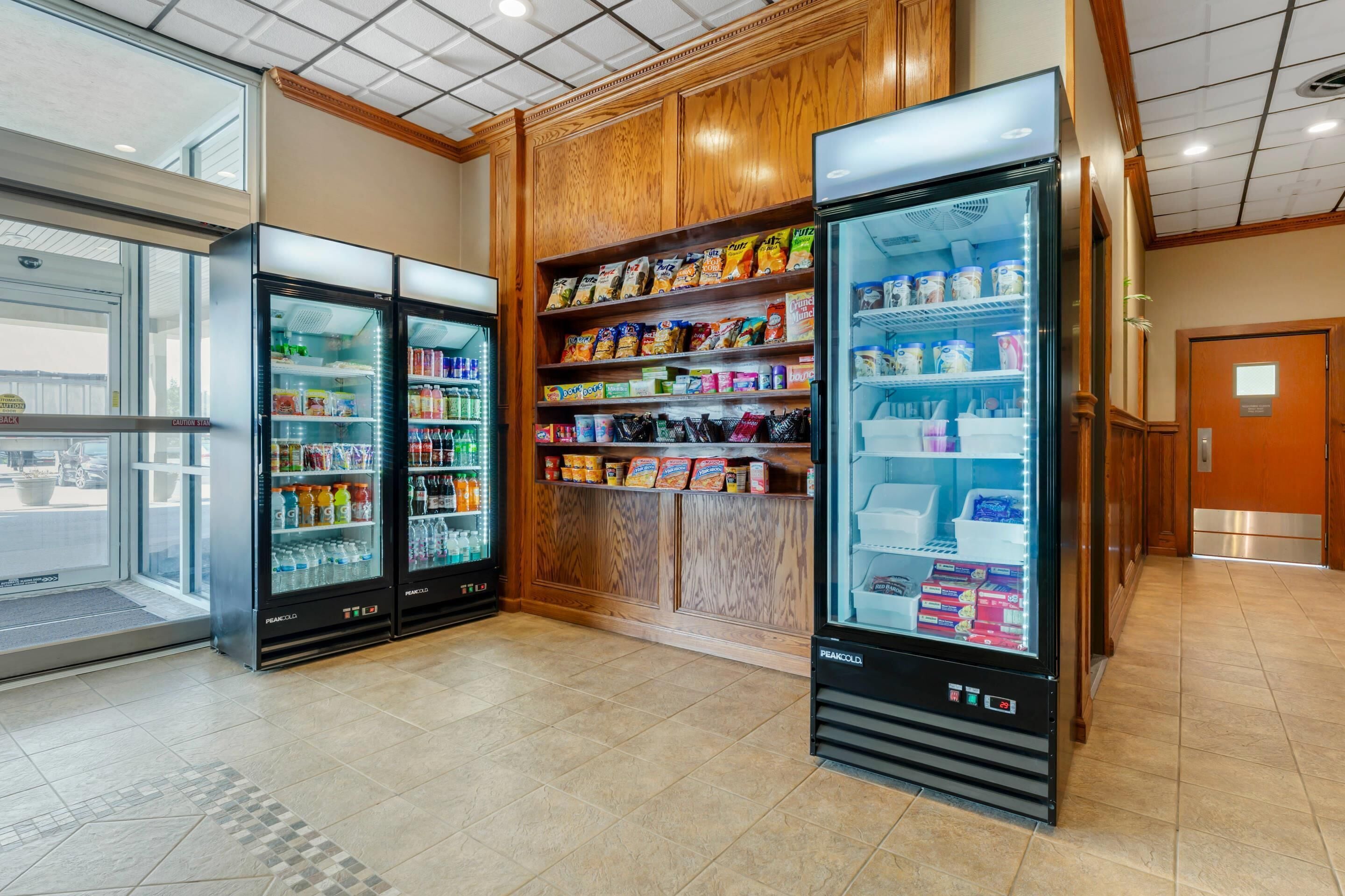 A hotel lobby with three refrigerated beverage dispensers, a snack bar with various packaged foods, and a wooden panel wall.