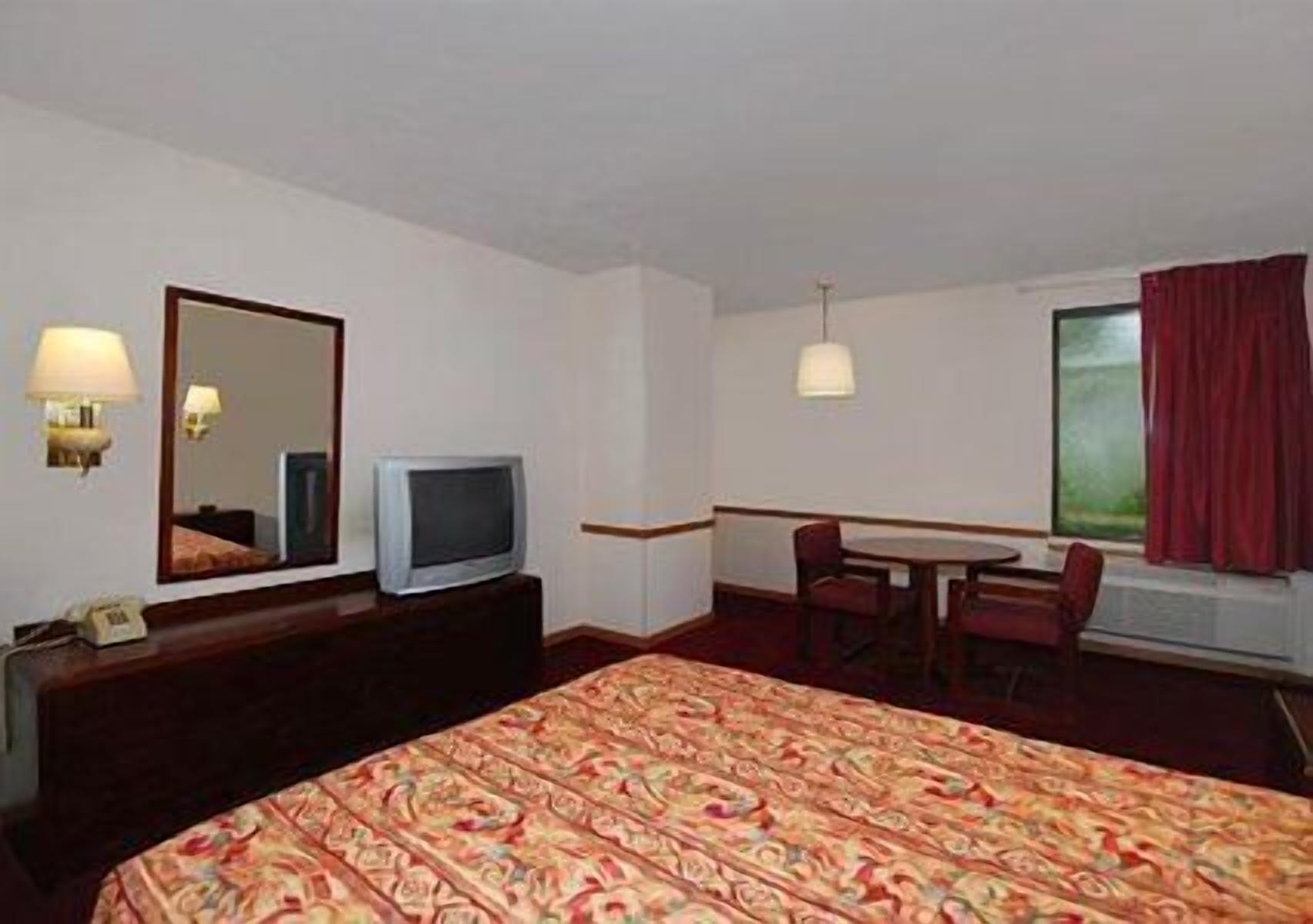Habitación de hotel con una cama, un televisor en una cómoda, un escritorio con dos sillas, una ventana con cortinas rojas y una lámpara fijada a la pared.