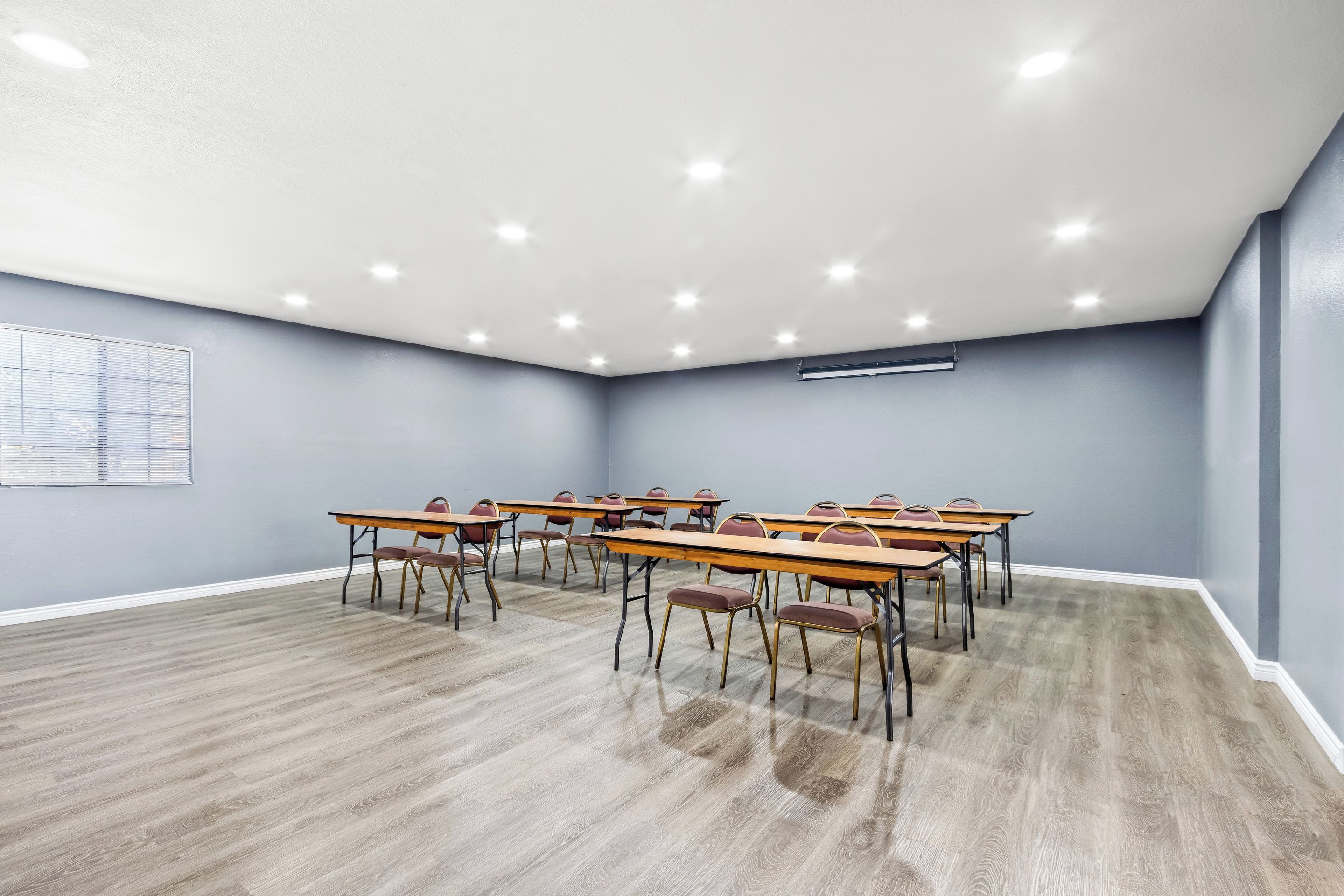 A conference room with long tables and chairs, recessed ceiling lights, and a window with blinds.
