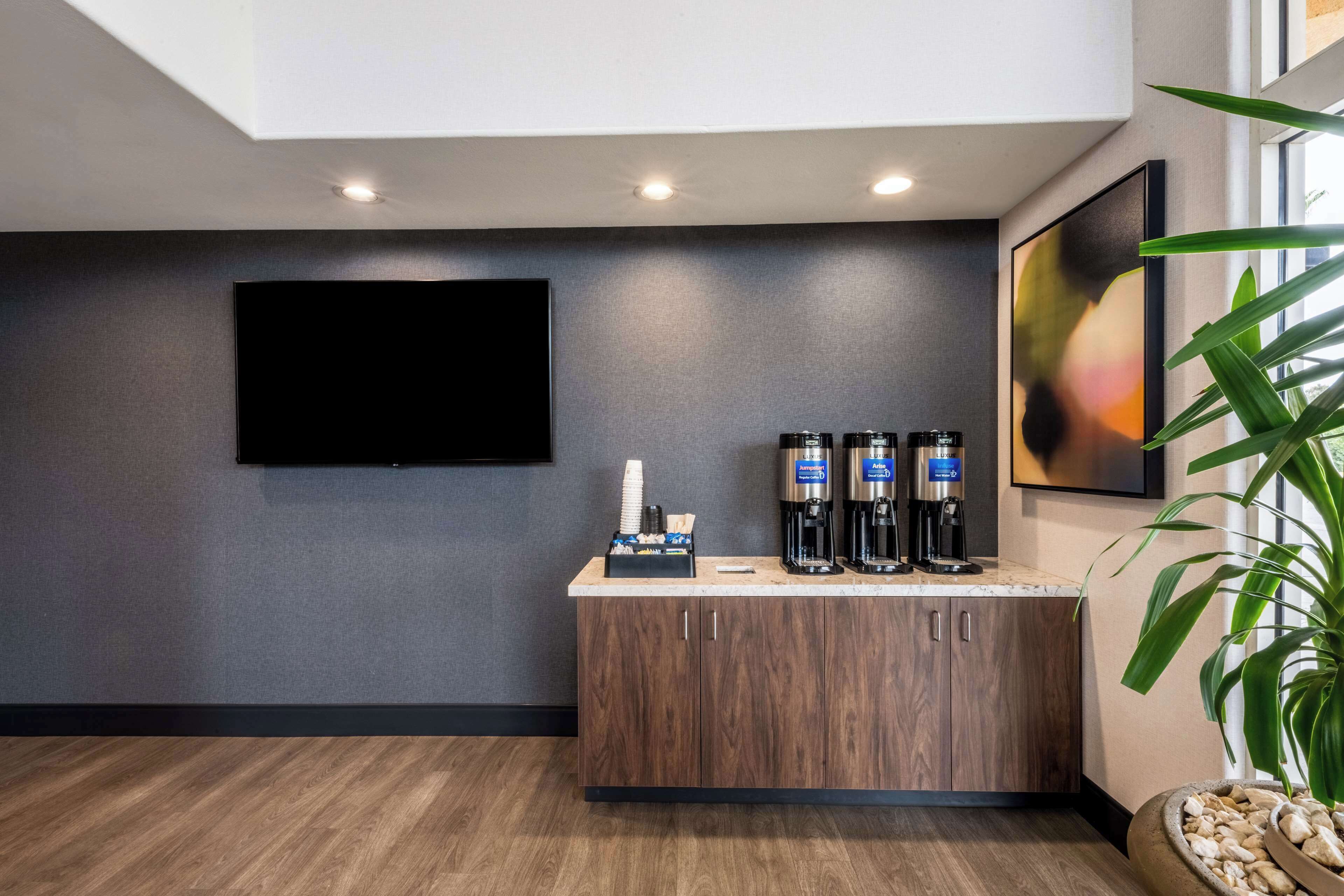 Un moderno vestíbulo de hotel con un televisor de pantalla plana montado en la pared, una estación de café con tres máquinas de café y una gran planta en maceta.