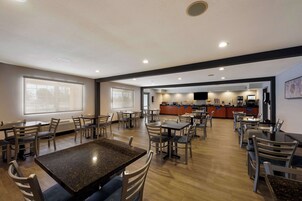 Un grand espace dédié à la salle à manger, avec plusieurs tables et chaises, une cuisine à l’arrière-plan et de grandes fenêtres qui laissent entrer la lumière naturelle.