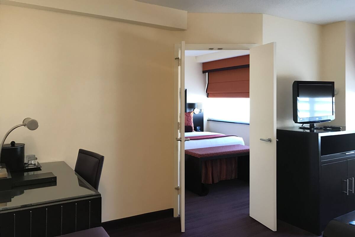 Quarto de hotel com cama, escrivaninha com luminária, televisão e guarda-roupa.