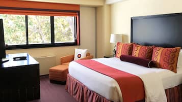 A hotel room with a large bed, a desk with a television, a window with a view of trees, and a nightstand with a lamp.