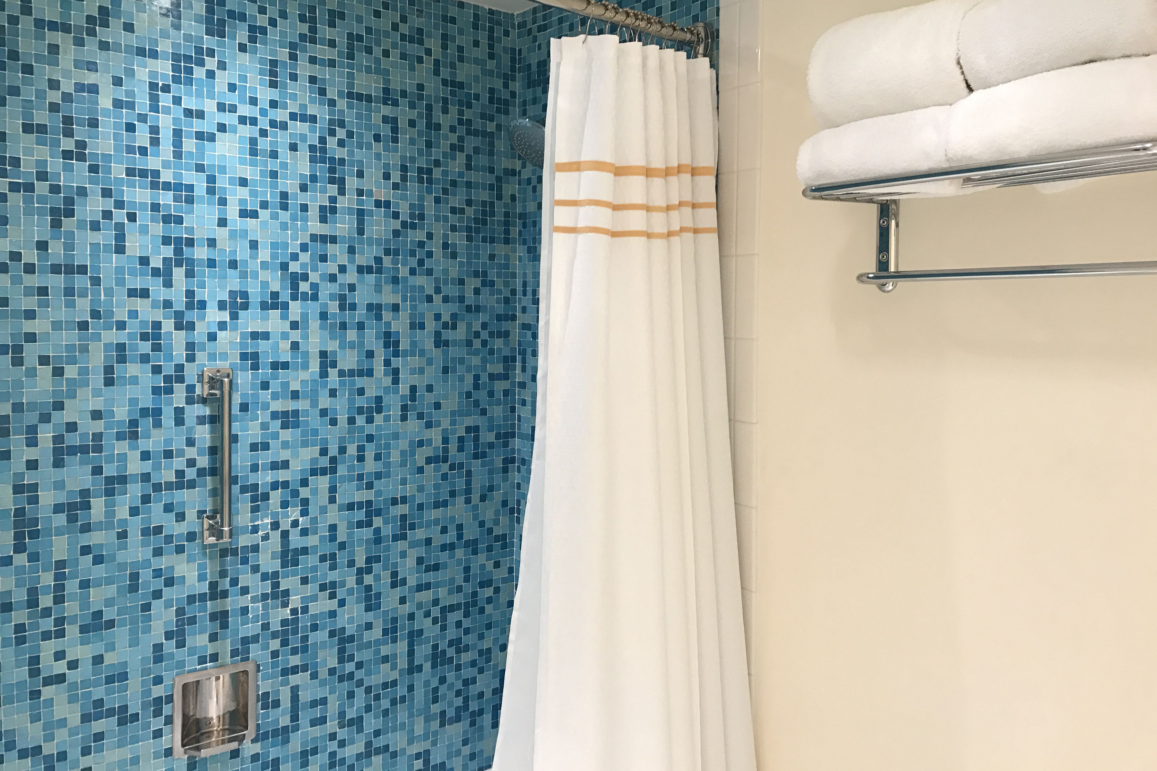 Un baño con pared de mosaico azul, cortina de ducha blanca con rayas anaranjadas y un perchero con toallas dobladas.