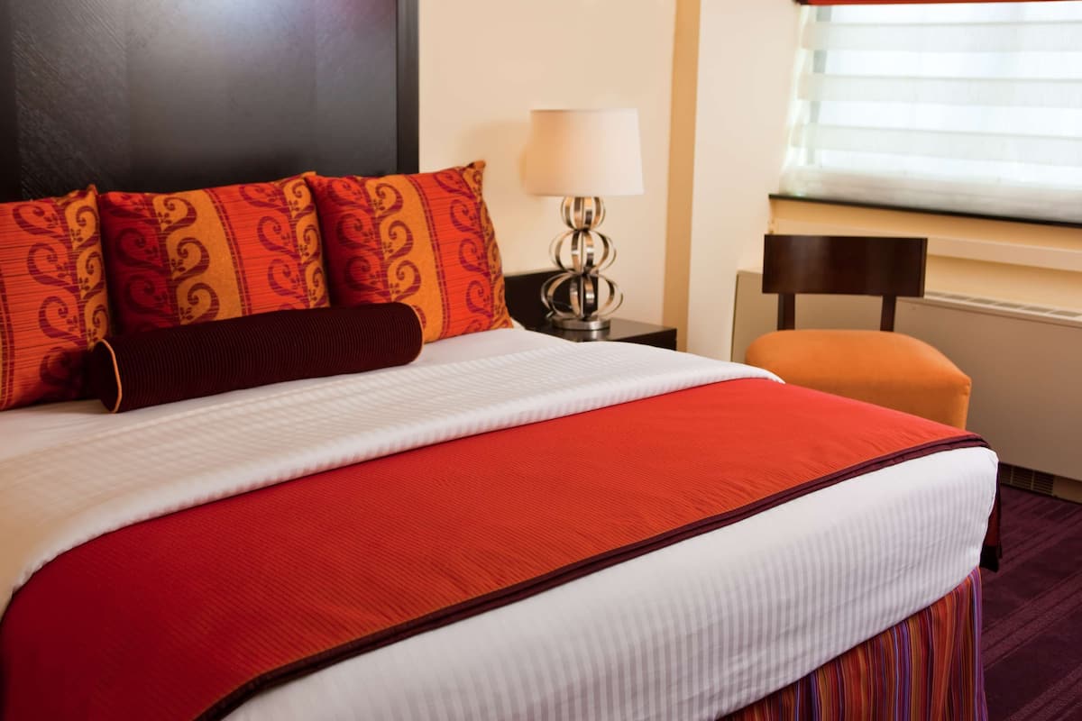 Cama bem arrumada com colcha vermelha e branca, várias almofadas com padrão vermelho e laranja, abajur e janela com persianas.