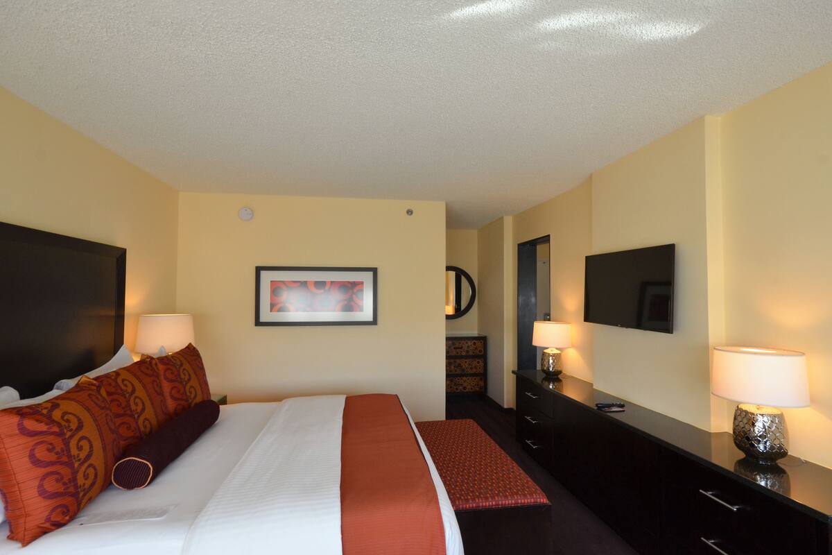 Quarto de hotel com uma cama, duas luminárias de cabeceira, uma televisão, cômoda e um quadro pendurado na parede.