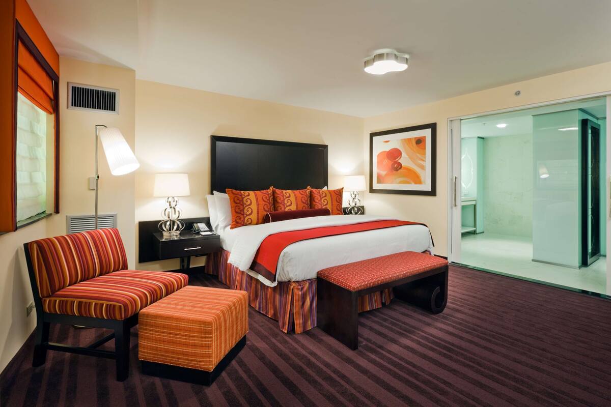 Quarto de hotel com uma cama grande, uma escrivaninha, uma cadeira, um banquinho e uma moldura de chuveiro de vidro, além de um quadro na parede.