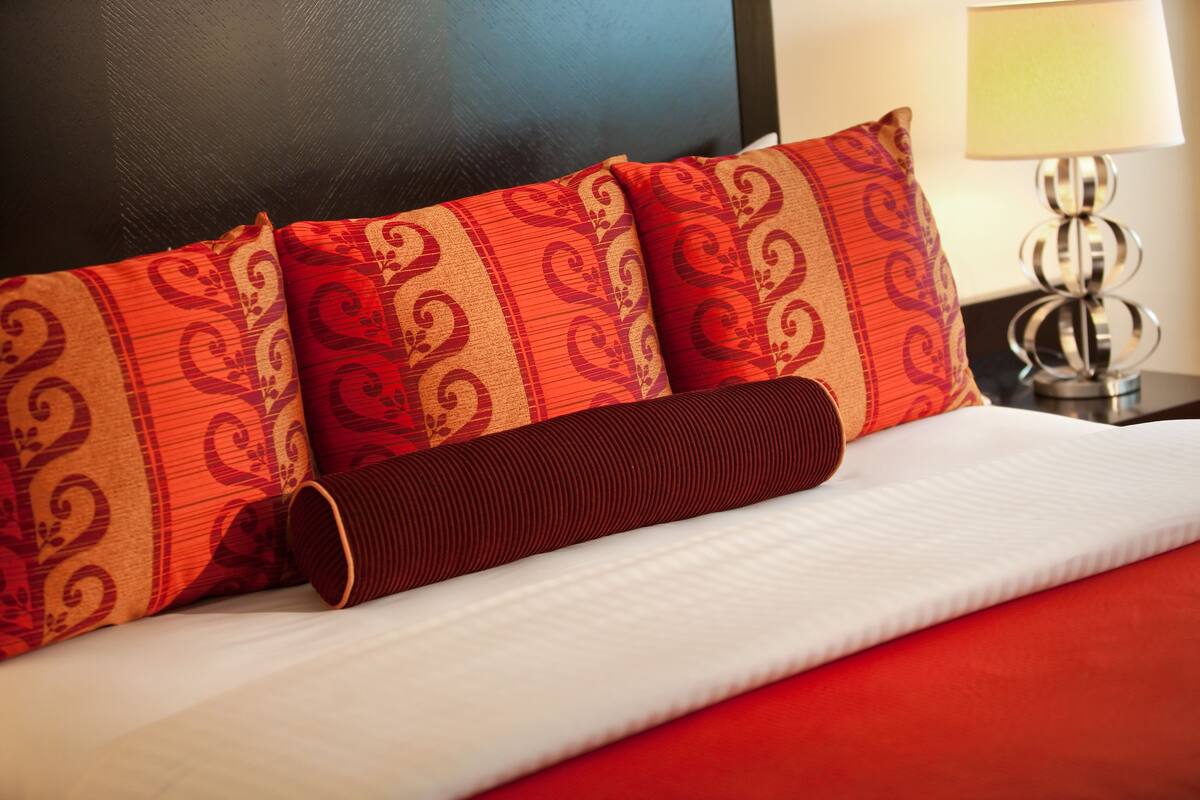 Uma cama com almofadas estampadas em vermelho e bege, um travesseiro cilíndrico marrom e um travesseiro branco.