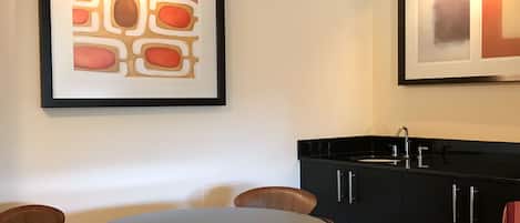 Un coin repas avec une table ronde et des chaises, un meuble noir avec un évier, et deux tableaux abstraits encadrés accrochés au mur.
