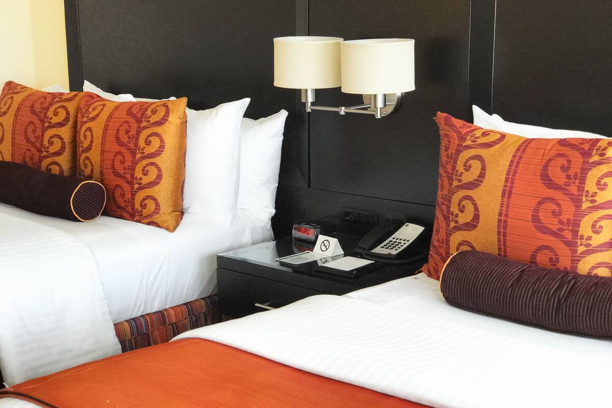 Quarto de hotel com duas camas, cada uma com roupa de cama branca e almofadas estampadas. Há uma mesa de cabeceira com telefone, relógio e placa indicando proibição de fumar.