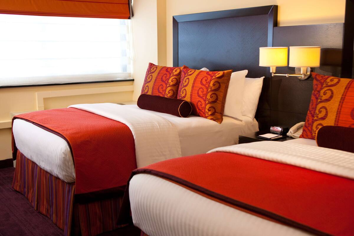 Quarto de hotel com duas camas, cada uma com roupa de cama vermelha e branca, cabeceira escura e mesa de cabeceira com abajur e telefone.