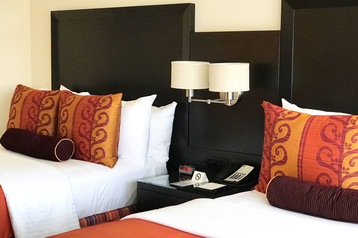 Quarto de hotel com duas camas, cada uma com almofadas estampadas, uma mesa de cabeceira com telefone e relógio, e luminárias fixadas na parede.
