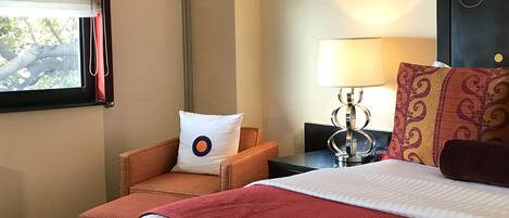 Habitación de hotel con cama, una silla, mesita de noche, lámpara y ventana con cortinas.